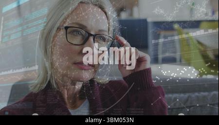 Woman adjusting black glasses while analyzing digital overlays on sofa wearing burgundy cardigan Stock Photo