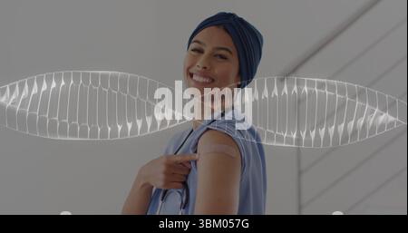 Smiling medical worker in scrubs and turban pointing to vaccine bandage at clinic hall, stethoscope Stock Photo