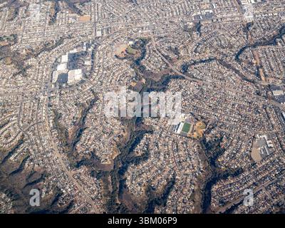 An aerial view shows a densely populated suburban area with winding streets and numerous houses, interspersed with patches of green space and trees, highlighting the urban planning and residential layout of the community. Stock Photo
