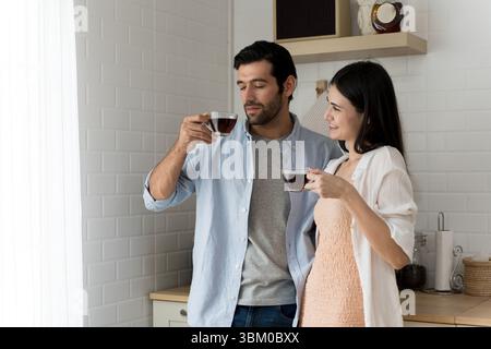 Young couple drinking coffee together in modern kitchen. Man and woman holding glass cups, sharing a peaceful morning moment with smiles. Cozy lifesty Stock Photo