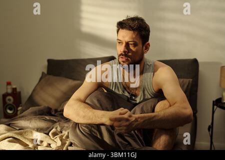Young adult man sitting on bed holding knees looking away with troubled expression suggesting family problems in domestic bedroom setting during daytime Stock Photo