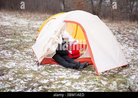 Two children sit closely inside a tent, wrapped in warm clothing, enjoying each other's company amid a snowy field on a cold winter day. Stock Photo