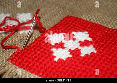 The detailed process of cross-stitch embroidery on burlap fabric. A metal embroidery needle is threaded with vibrant red thread. Stock Photo