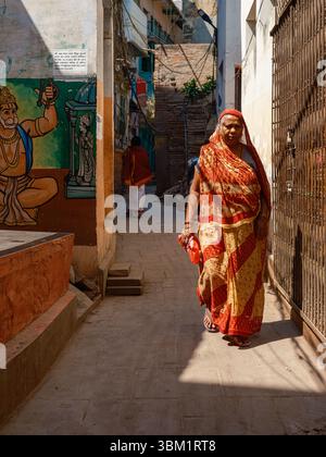 India, Varanasi, 16 Mar 2025. Woman in a red-and-gold sari walks through a sunlit alley past a Hanuman mural, framed by shuttered doors and narrow tem Stock Photo