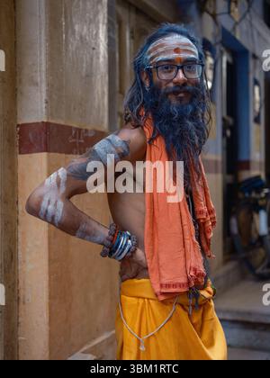 India, Varanasi, 16 Mar 2025. Hindu sadhu with painted forehead and arms, wearing saffron robes and beaded bracelets, stands in a narrow alley near th Stock Photo