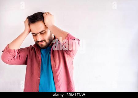 A young man with a beard, wearing a pink shirt over a blue t-shirt, stands against a plain white background, hands on his head, expressing stress or e Stock Photo