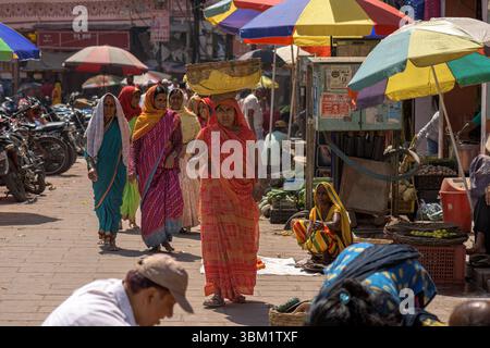 India, Varanasi, 16 Mar 2025. Women in colourful saris walk through a bustling market street lined with fruit stalls and rainbow-shaded umbrellas unde Stock Photo