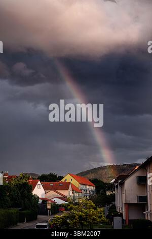 A vibrant rainbow arcs across the sky above a small residential town with red-tiled rooftops. Dark storm clouds still loom overhead while sunlight bre Stock Photo