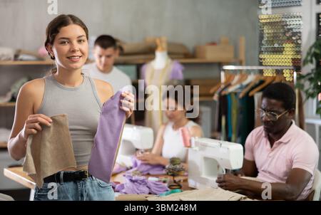 Young female participant present fabric billet, others sewing and working in background Stock Photo
