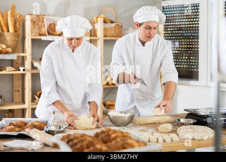 Focused female and guy baker in white uniform working at bakery, kneading and cutting dough into pieces Stock Photo