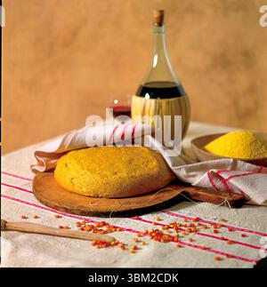 Round loaf of corn bread on a wooden cutting board, polenta flour in a bowl, a fiasco of red wine, a glass of wine and some corn kernels scattered on Stock Photo