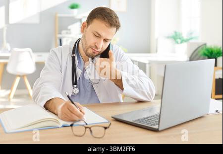 Male serious doctor, focused man talking to patient on mobile phone in office, using laptop  Stock Photo