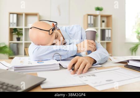 Overworked tired young man putting head on table in office with documents holding cup of coffee. Stock Photo