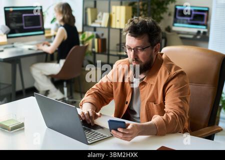 Caucasian Young Adult Man Developing Neural Network Using Laptop and Smartphone Stock Photo