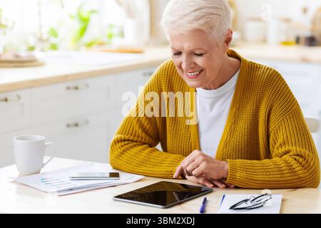 Joyful grey-haired senior woman using digital tablet in kitchen Stock Photo
