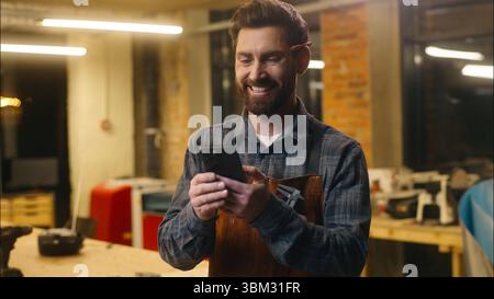 Happy Caucasian man carpenter using mobile phone in workshop wood master craftsman swiping screen device smiling artist male architect order hardwood Stock Photo