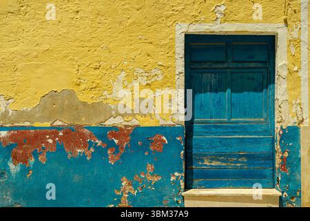 An old, weathered wooden door, painted in a vibrant turquoise blue, is set into a wall with peeling yellow and blue paint in Ciutadella, Spain. Stock Photo