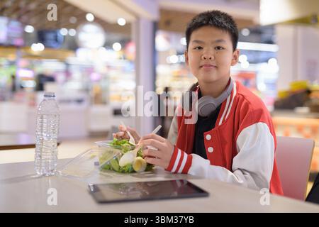 Handsome Teen Thai boy wearing headphones eating salad in cafeteria Stock Photo