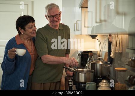 Elderly couple sharing moment while cooking in modern kitchen. Senior man stirring pot while woman holding cup, both smiling and enjoying time together Stock Photo