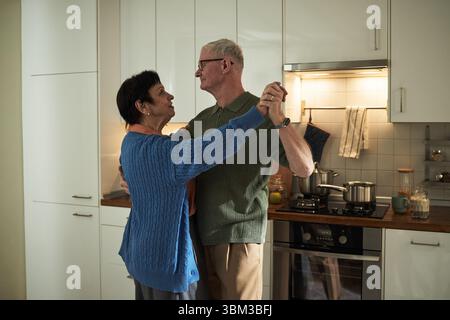 Elderly couple dancing in their modern kitchen, sharing a joyful moment, illuminated by overhead lighting and surrounded by contemporary kitchen appliances, at home Stock Photo