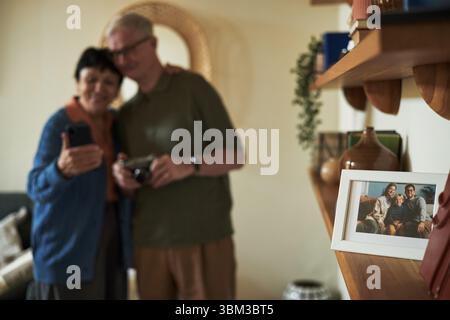 Senior couple standing closely, smiling, and posing with smartphone in cozy living room. Background includes framed photo on shelf and warm home decor creating a welcoming environment Stock Photo