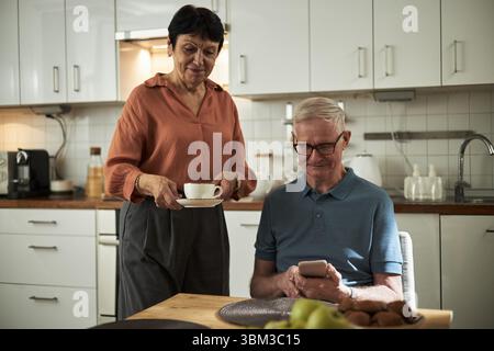 Elderly couple sitting in modern kitchen, man using smartphone while woman smiling and holding cup of tea. Atmosphere of warmth and tranquility surrounding them Stock Photo