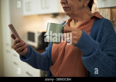 Senior woman standing in kitchen holding a mug while using smartphone, wearing a blue sweater. Appliances visible in the background Stock Photo