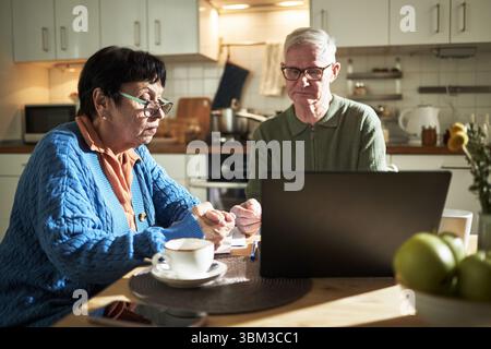 Elderly couple sitting at kitchen table, engrossed in using laptop and taking notes, surrounded by coffee cup and notepad, natural light illuminating the scene Stock Photo