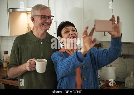 Smiling elderly couple taking selfie in modern kitchen while holding coffee mug, capturing cheerful moment and engagement in selfie activity, adding warmth to home setting Stock Photo