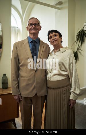 Portrait of beautiful elderly couple standing in a well-decorated living room, smiling gently. Light filtering through windows creates a pleasant atmosphere, showcasing their elegant clothing Stock Photo