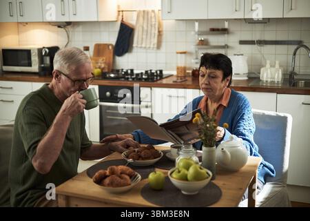 Elderly couple sitting at table having coffee and reading magazine in cozy kitchen. Seated on chairs, table adorned with fresh fruit, muffins, vase with yellow flowers creating welcoming atmosphere Stock Photo