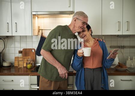 Elderly couple standing in kitchen, smiling while having morning coffee and using smartphone. Senior lady holding mug, senior man embracing affectionately Stock Photo