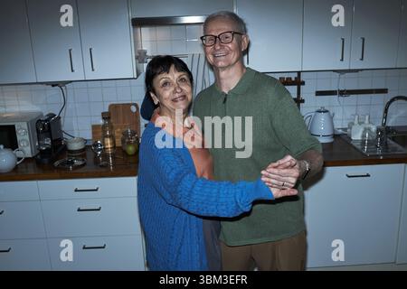 Elderly couple smiling and holding hands while dancing in cozy kitchen. Warm and joyful atmosphere with kitchen appliances and wooden countertop visible Stock Photo