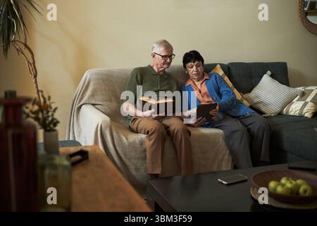 Senior couple sitting on a couch and reading books, seemingly engaged in their individual activities, creating a cozy and intimate atmosphere in the living room Stock Photo