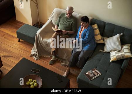 Elderly couple sitting on cozy sofa in living room, reading book and novel on digital tablet, spending peaceful time together in relaxed atmosphere Stock Photo