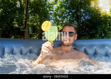 Bavaria, Germany - June 24, 2025: Concept for refreshment at home: A man cools off in the pool with a cold drink and enjoys the summer. The picture stands for cooling off and relaxing on a hot day in your own garden. *** Konzept für Erfrischung zuhause: Ein Mann kühlt sich im Pool mit einem kalten Getränk und genießt den Sommer. Das Bild steht für Abkühlung und Entspannung an einem heißen Tag im eigenen Garten. Stock Photo
