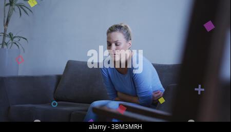 Leaning woman clutching abdomen, sitting on gray sofa in lounge, with plant and floating shapes Stock Photo