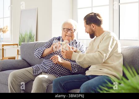Senior father sitting on sofa at home with his adult son and using smartphone. Stock Photo