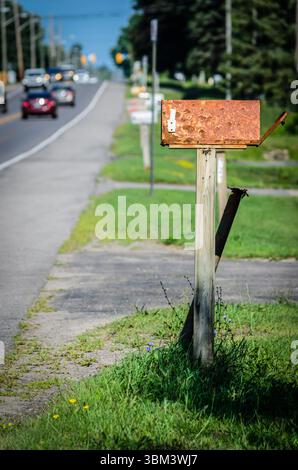 Rusty rural mailbox in front of a row of out of focus mail boxes next to a busy road in the summer. Stock Photo