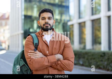 A young, bearded man stands confidently with arms crossed, wearing a button-down shirt, and a backpack. The background features a modern building. Stock Photo