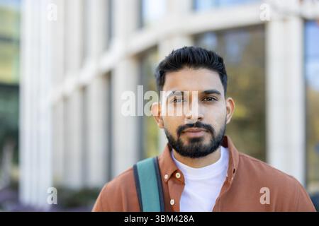 A young man with a beard and a backpack stands in front of a building on a sunny day. Stock Photo