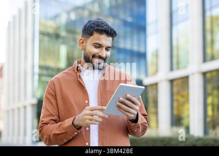 A smiling man with a beard uses a tablet outside a modern building on a sunny day, looking at the screen. Stock Photo