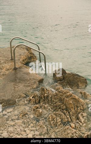 A rocky shoreline with a simple metal ladder leading into the calm, clear sea. Pebbles are visible beneath the water, and gentle waves. Stock Photo