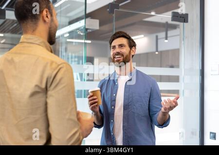 Two men are chatting in an office, one holding a coffee cup and gesturing while the other listens. Stock Photo