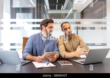 Two diverse businessmen collaborate in a modern office, discussing project details with laptops and documents. Stock Photo