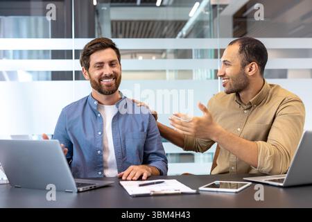 Two diverse male colleagues smile and collaborate in a modern office setting, using laptops and other digital devices. Stock Photo