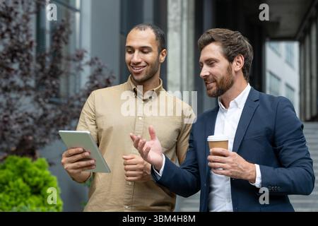 Two businessmen reviewing a tablet outdoors near an office building, discussing and drinking coffee, with natural expressions and smiles. Stock Photo