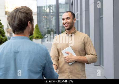 Two businessmen shaking hands in front of a modern building. One man holds a tablet and smiles. Stock Photo