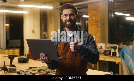 Cheerful Caucasian man carpenter typing laptop showing thumb up sign in workshop wood master male worker artist using Internet delivery app online Stock Photo