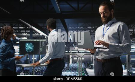 Computer scientists on data center industrial platform analyzing performance outputs on laptop. Server farm IT experts collaborating on finetuning rigs configurations, Stock Photo